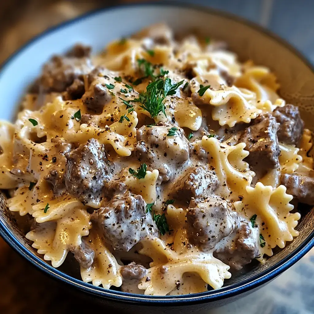 A bowl of creamy pasta with bowtie noodles, chunks of beef, and parsley, sprinkled with black pepper.