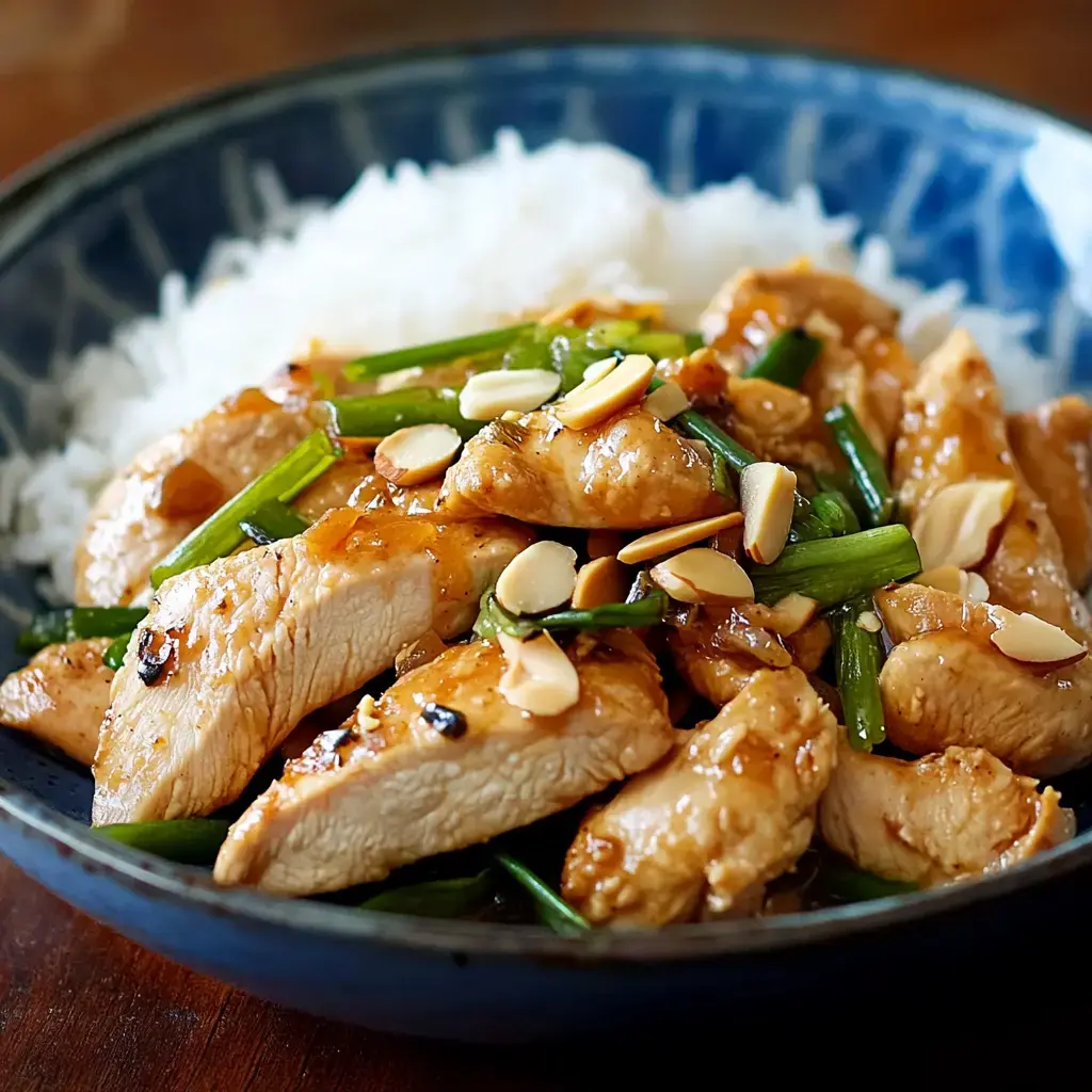 A bowl of sliced chicken in a savory sauce, garnished with green onions and almonds, served over a bed of white rice.