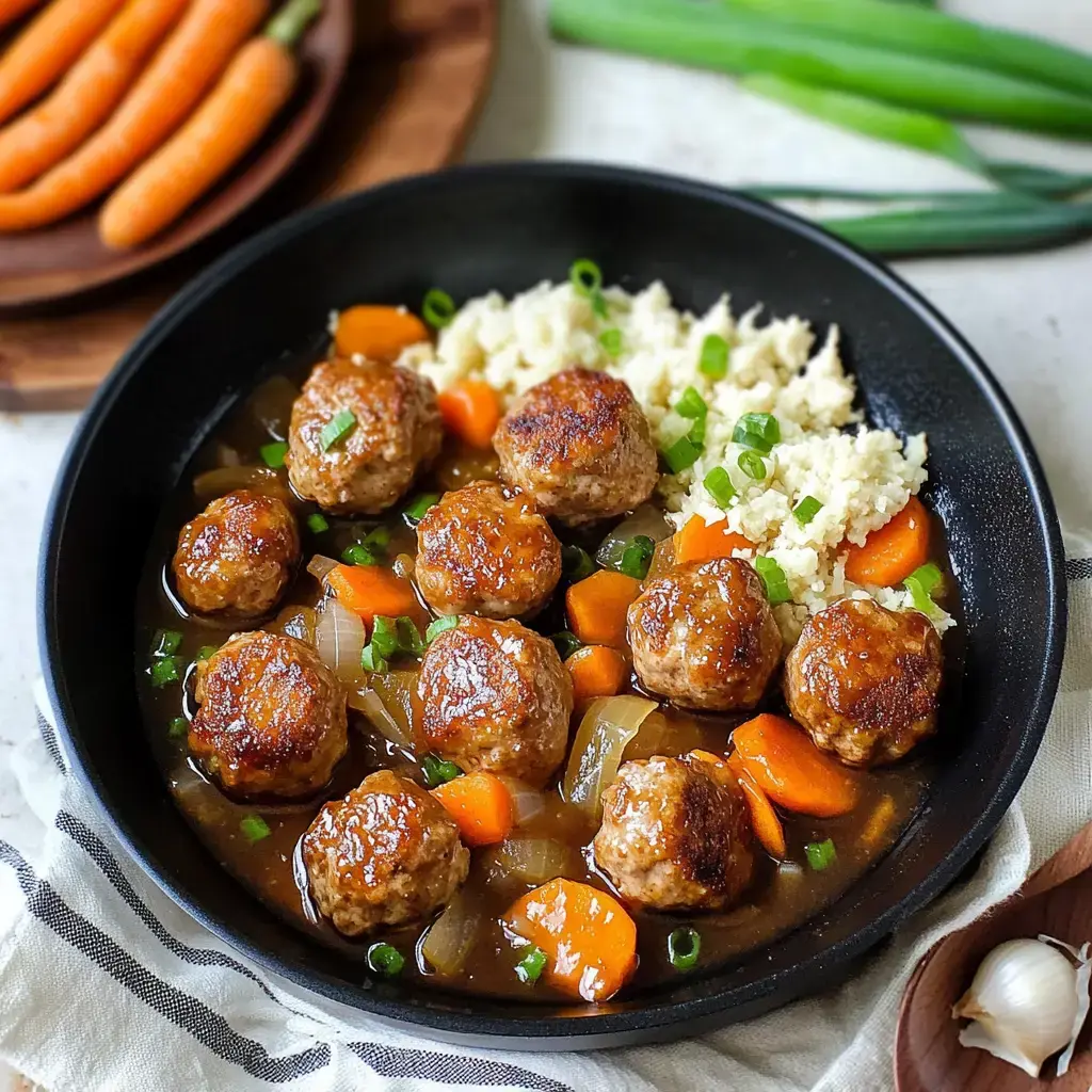 A black bowl contains meatballs in a savory sauce, surrounded by carrots and onions, served with a side of cauliflower rice and topped with green onions, with fresh carrots and garlic in the background.