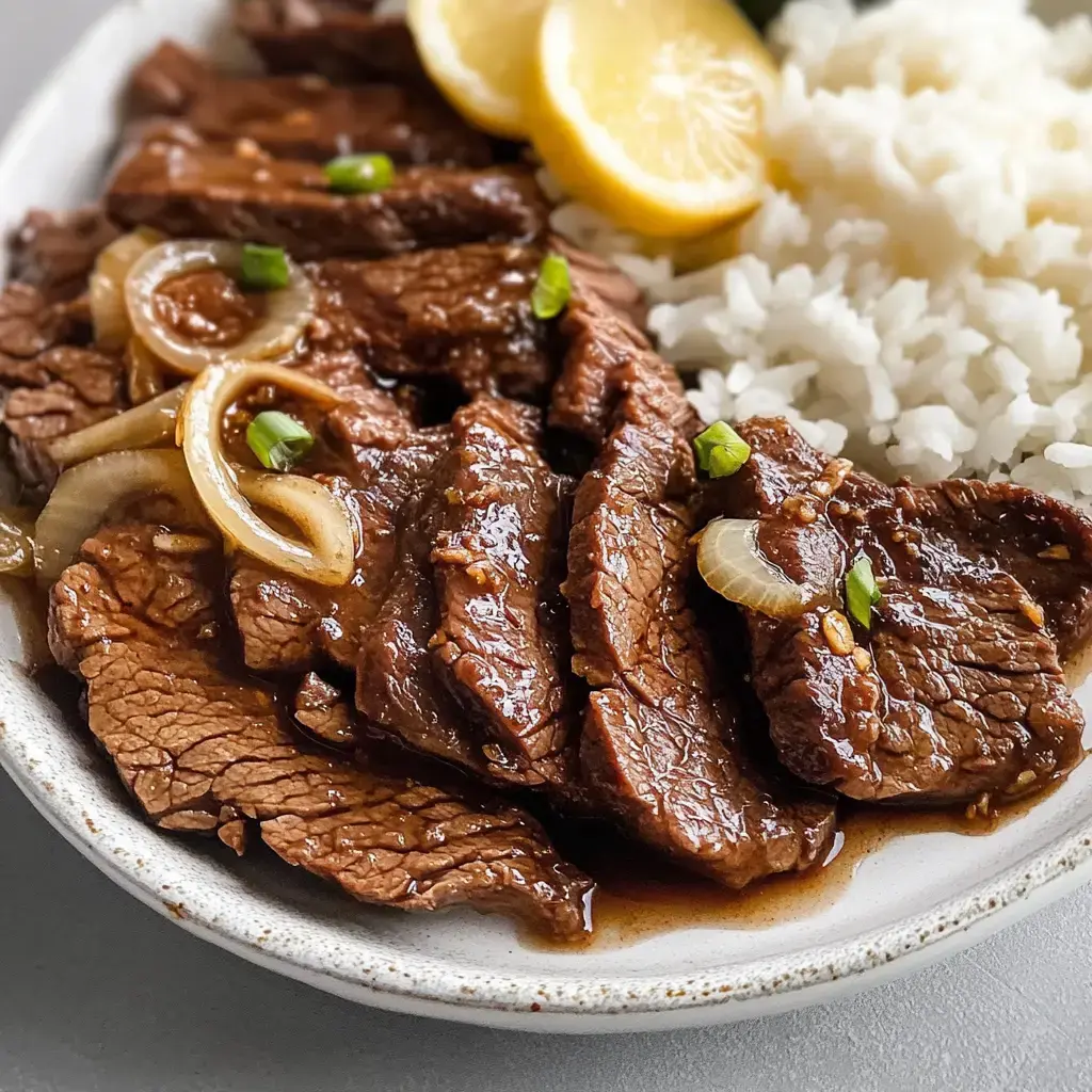 A plate of sliced beef in a savory sauce is accompanied by white rice and lemon wedges.