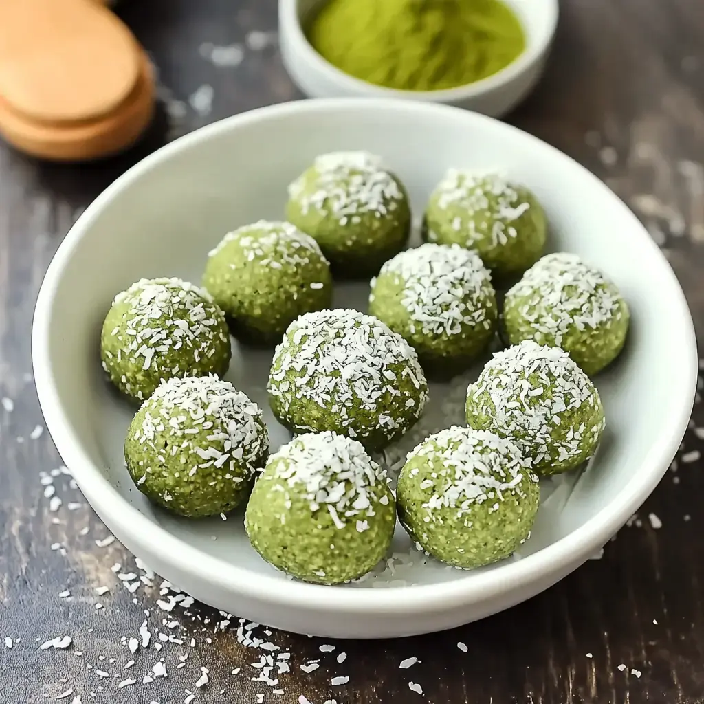 A white bowl holds several green energy balls covered in coconut flakes, placed on a dark wooden surface.