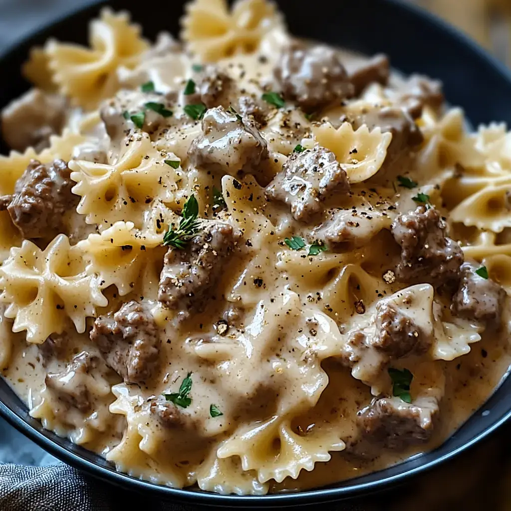 A bowl of farfalle pasta mixed with creamy sauce and pieces of beef, garnished with parsley and black pepper.