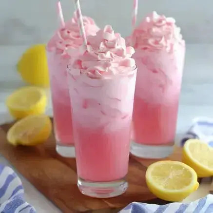 Three tall glasses filled with pink swirled beverages topped with whipped cream, accompanied by fresh lemon halves on a wooden cutting board.