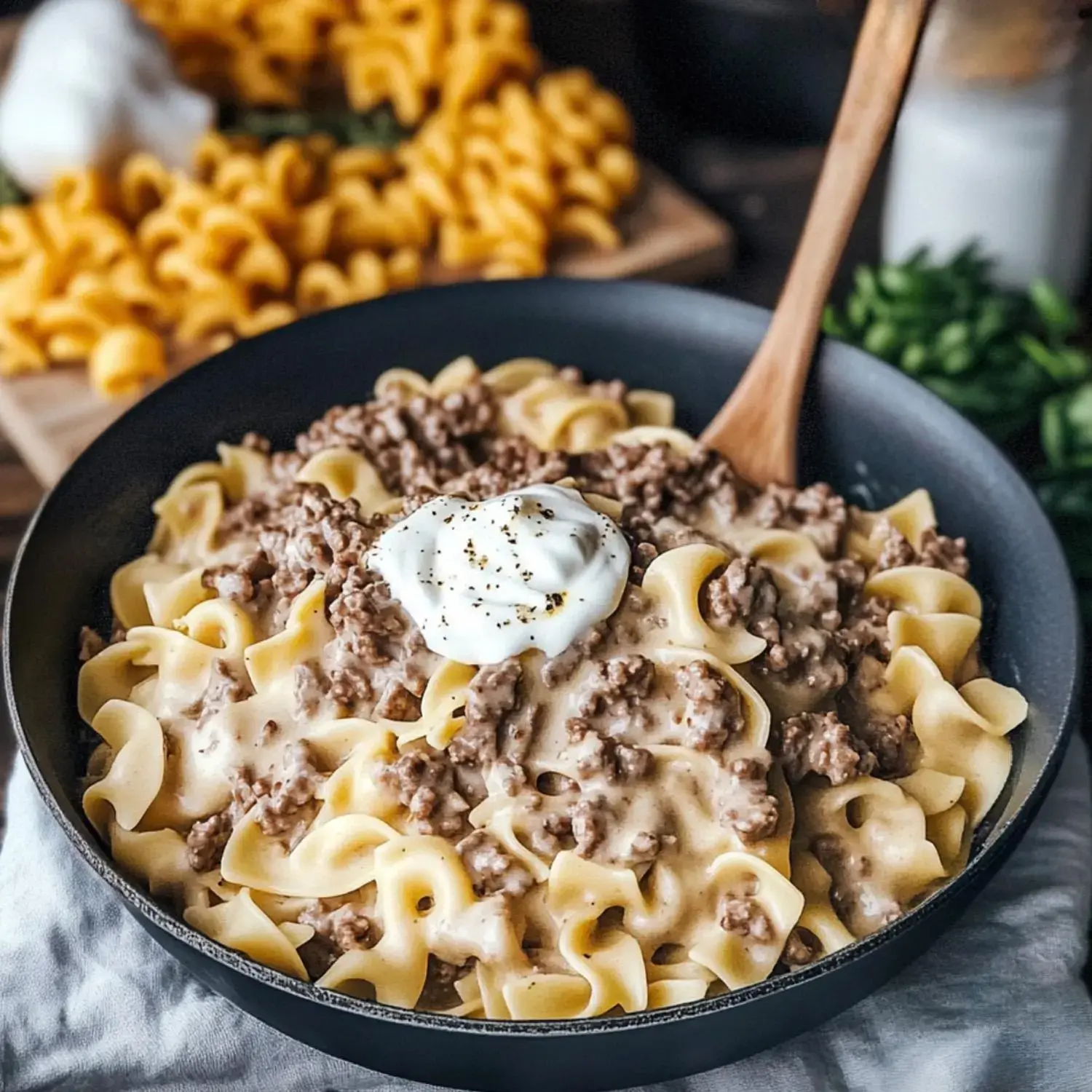 A serving of egg noodles topped with creamy ground beef and a dollop of sour cream in a black bowl, with uncooked pasta in the background.