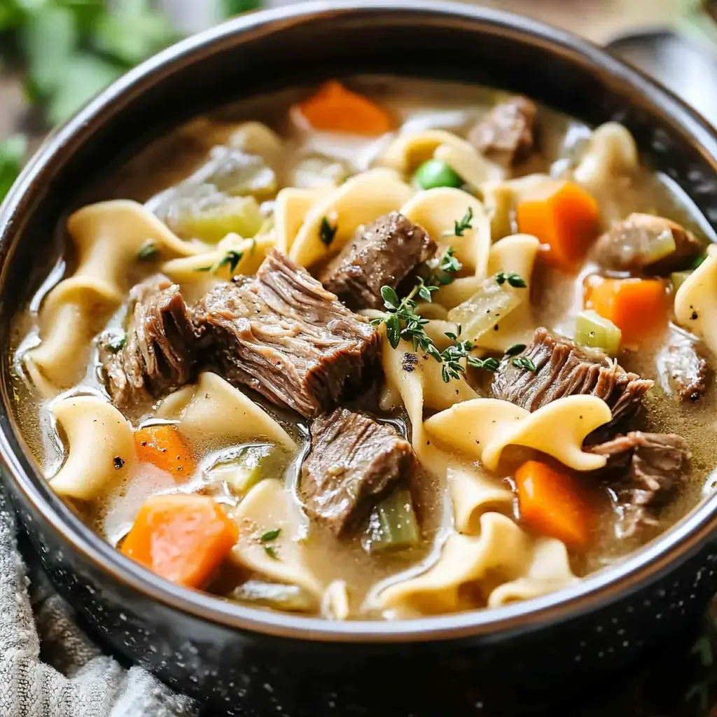 A close-up of a hearty beef noodle soup containing chunks of beef, pasta, carrots, celery, and herbs in a dark bowl.