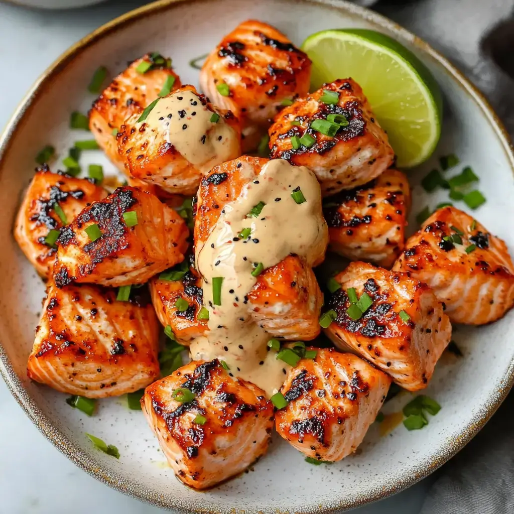 A plate of grilled salmon cubes garnished with green onions and drizzled with a creamy sauce, accompanied by a lime wedge.