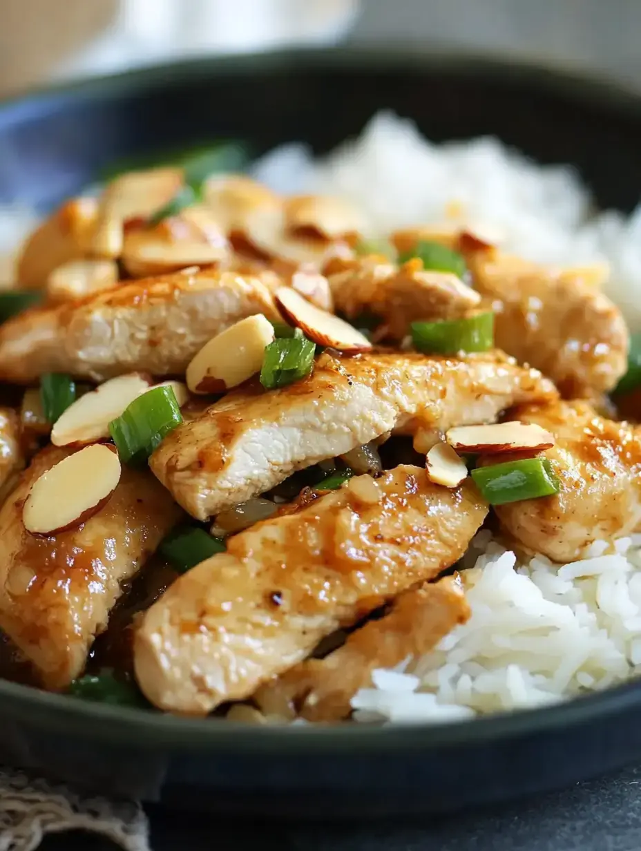 A close-up of sliced chicken breast topped with almonds and green onions served over fluffy white rice in a dark bowl.