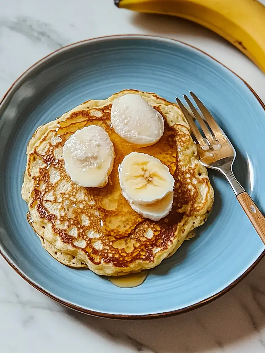 A plate of pancakes topped with banana slices and syrup, beside a fork and a partially visible banana.