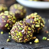 A close-up image of chocolate-covered truffles coated with crushed pistachios on a dark surface.