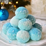 A decorative stack of blue candy truffles coated with sparkling sugar sits on a white plate, with a blue ornament and a softly glowing background.