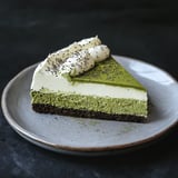 A slice of layered matcha cheesecake with a creamy top, adorned with whipped cream and sprinkled with poppy seeds, on a gray plate.