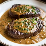 A plate of two grilled steaks topped with creamy sauce and garnished with chopped green herbs.