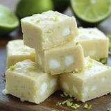 A stack of lime-flavored fudge squares, dotted with white bits and garnished with lime zest, against a backdrop of fresh limes.