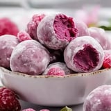 A close-up of a bowl filled with pink, powdered frozen dessert balls, some with bites taken out, surrounded by fresh raspberries.