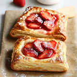 Two golden-brown pastry rectangles filled with strawberries and topped with powdered sugar, placed on parchment paper.