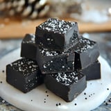 A stack of dark chocolate fudge pieces topped with coarse salt, displayed on a white marble surface.