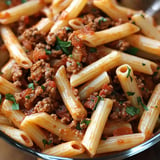 A close-up of penne pasta mixed with ground meat and a rich tomato sauce, garnished with chopped parsley.