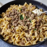 A close-up view of a bowl of creamy beef stroganoff served over egg noodles, garnished with parsley and a dollop of sour cream.