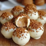 A close-up of white chocolate-covered truffles topped with crushed cookies, with one truffle cut open to reveal a creamy filling.