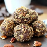 A close-up image of chocolate-covered truffles coated in crushed nuts, arranged on a wooden surface.