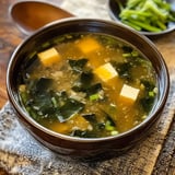 A bowl of green seaweed soup with tofu cubes and chopped green onions, placed on a textured cloth.