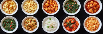 A variety of ten dishes, including pasta, vegetables, rice, and protein, arranged in white bowls on a dark surface.